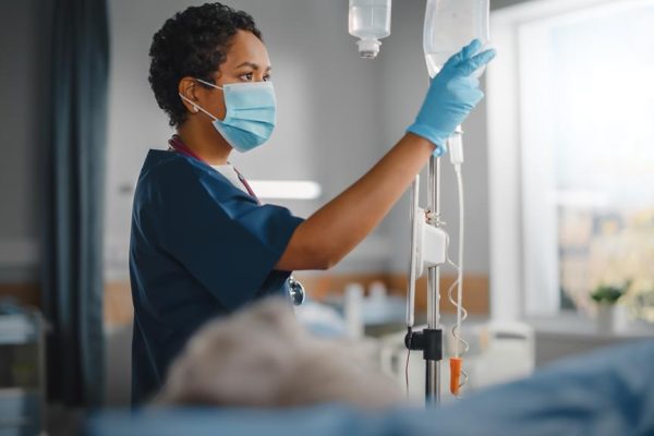 Hospital Ward: Professional Black Head Nurse Wearing Face Mask Does Checkup of Patient's Vitals, Checking Heart Rate Computer, Intravenous or Iv Fluids Drip Bag. Caring Nurse Monitors Person Recovery